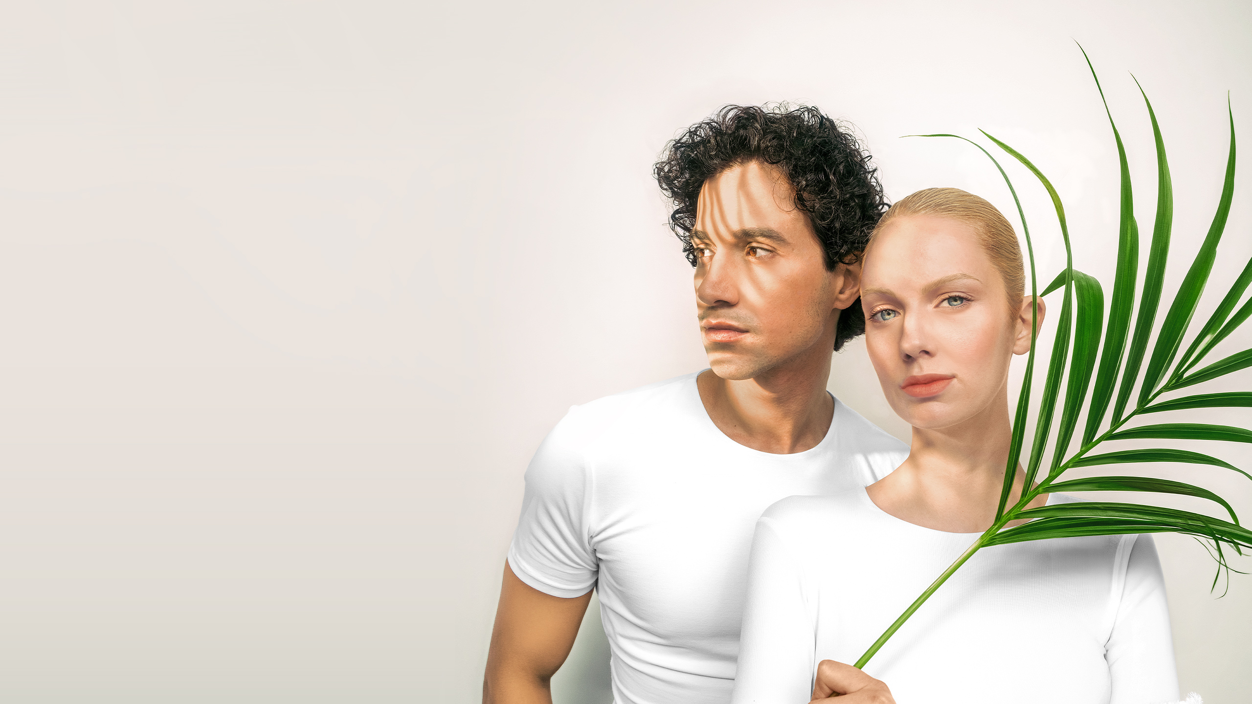 Two people dressed in white against a light background, partially framed by a green palm leaf.